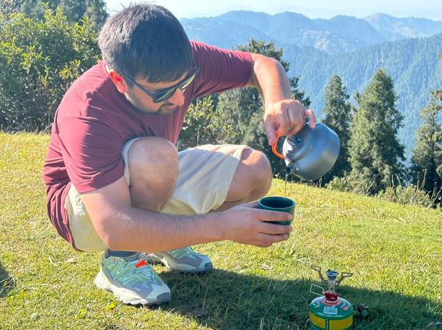 Backpacker using a compact backpacking stove to boil water on a grassy mountainside with scenic forested hills in the background.
