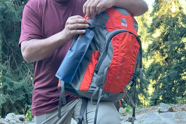Placing a collapsible water bottle into the side pocket of a red and gray hiking backpack.
