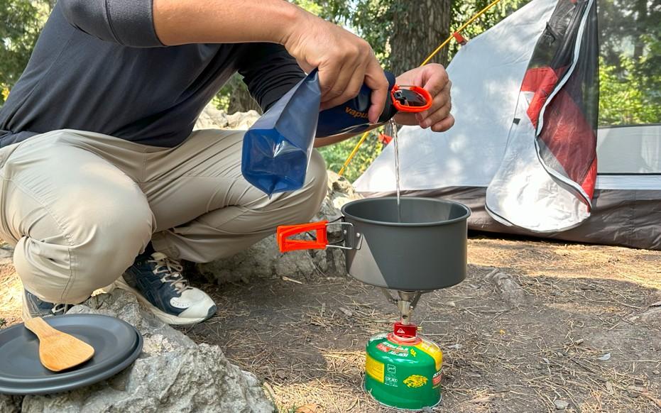 Camper pouring water into a pot on a backpacking stove at a forest campsite with tent setup nearby.