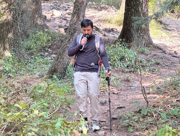 A hiker walks through a forest trail while drinking from a hydration bladder tube. He wears a black long-sleeve shirt, light hiking pants, and carries trekking poles and a backpack. Trees and foliage surround the rocky path.